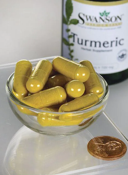 A small glass bowl filled with Swanson's Turmeric 720 mg capsules, known for curcuminoids that support joints, sits next to a U.S. penny on a clear surface. In the background, a bottle marked "Turmeric" completes the scene.