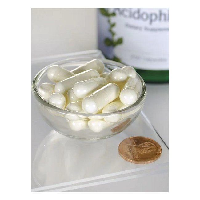 A small Swanson glass bowl with white capsules next to a penny highlights their size, suggesting digestive health benefits. A blurred Lactobacillus Acidophilus - 250 capsules bottle can be seen in the background.